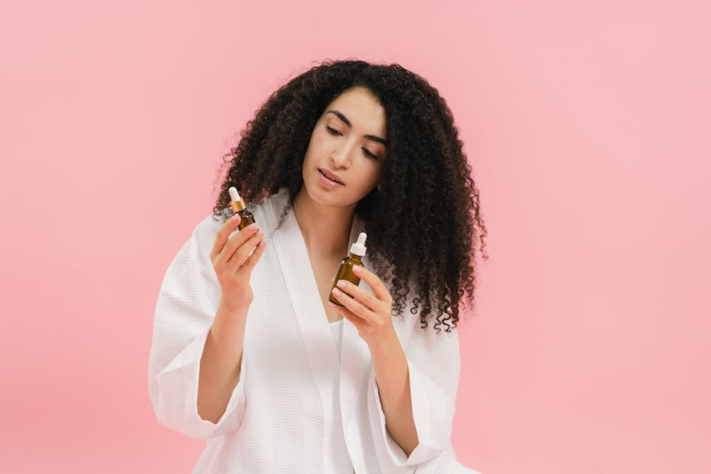 A woman with curly hair in a white robe examines two cosmetic oil bottles against a pink background.